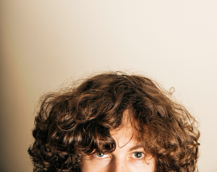 Close-up of a person with thick, curly brown hair and green eyes against a beige background as the page banner