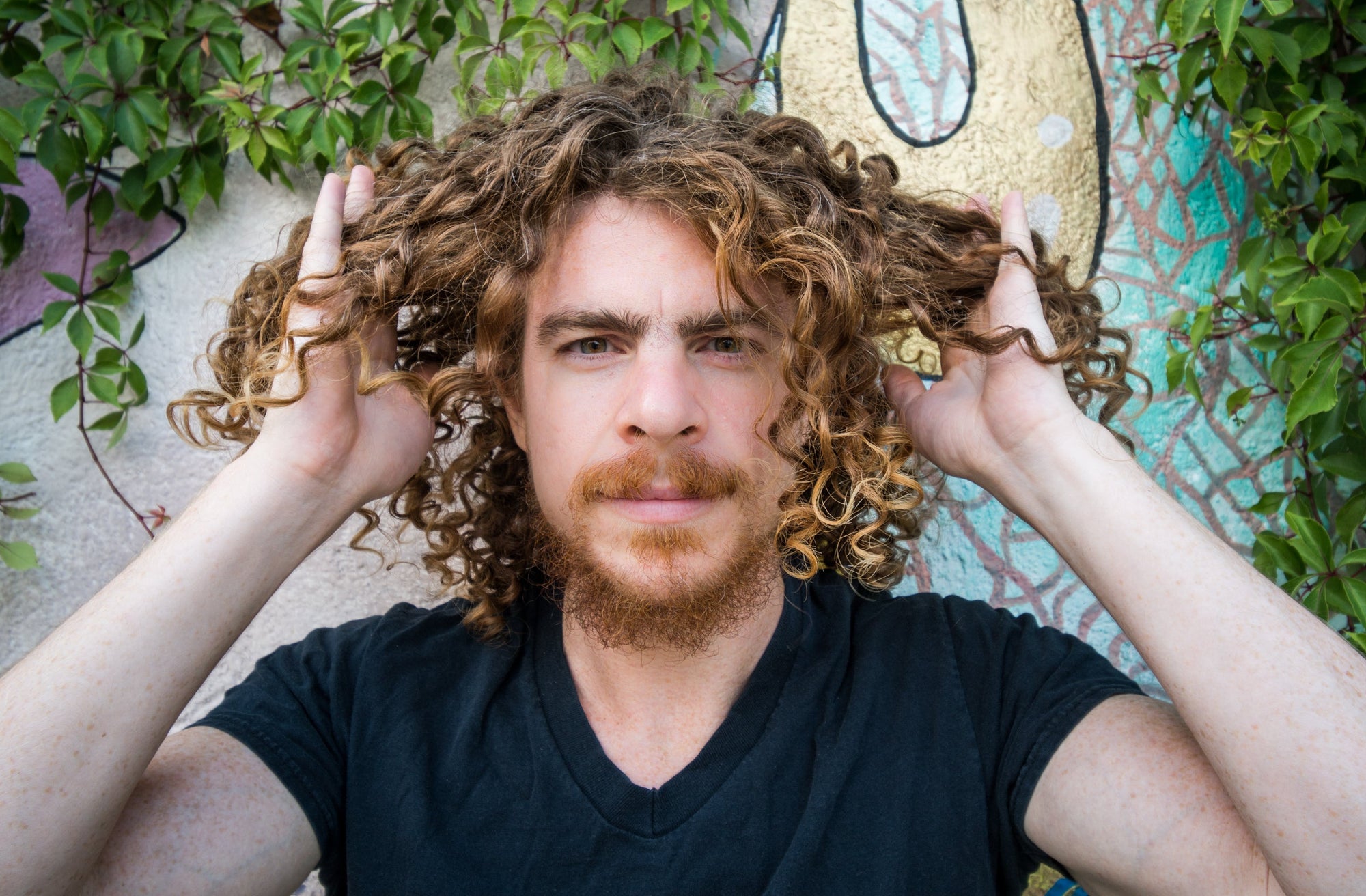 Man with curly hair holding his hair out to the sides in front of a colorful wall mural
