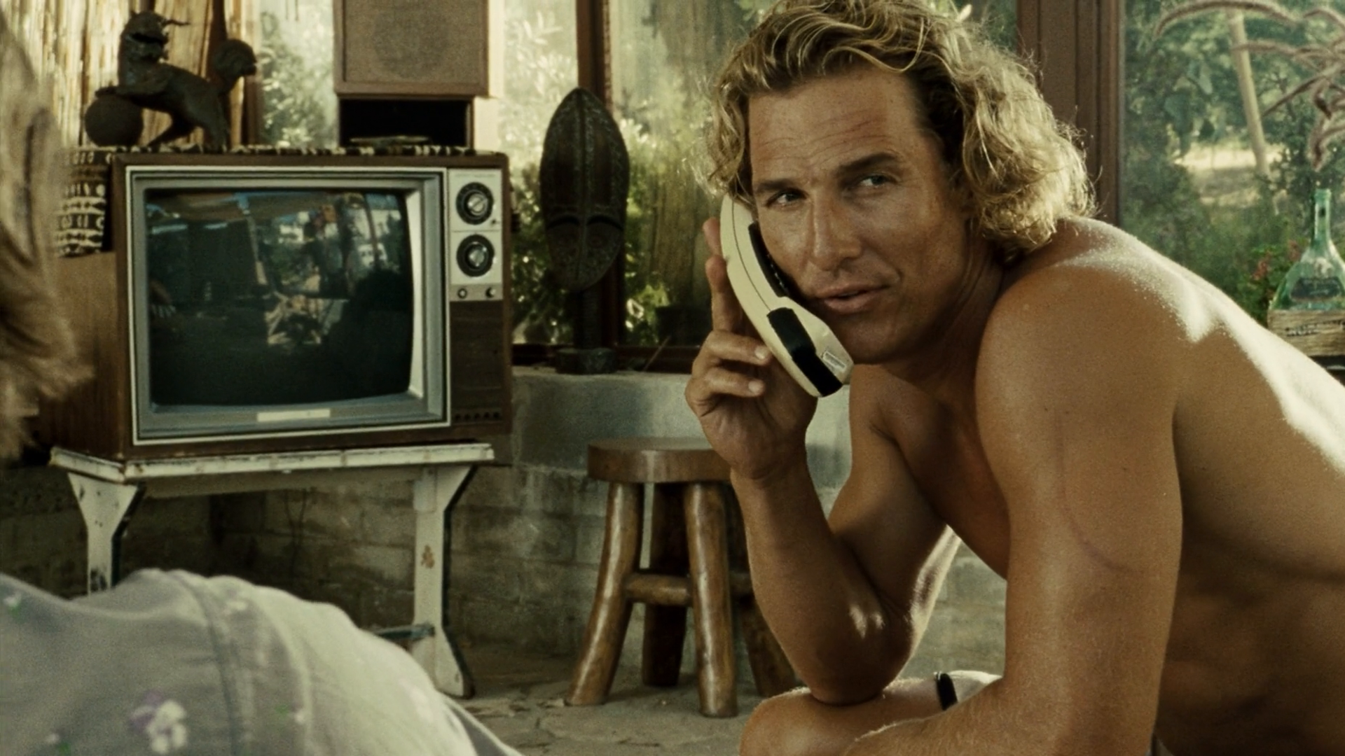 Shirtless man with sun-bleached surfer hair talking on a corded phone in a rustic surf-house with an old television behind him