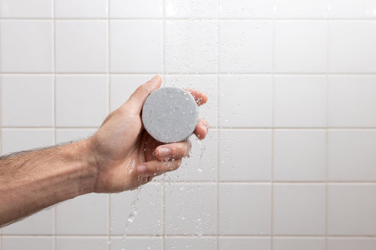 Hand holding a round gray shampoo bar under running water in a tiled shower.