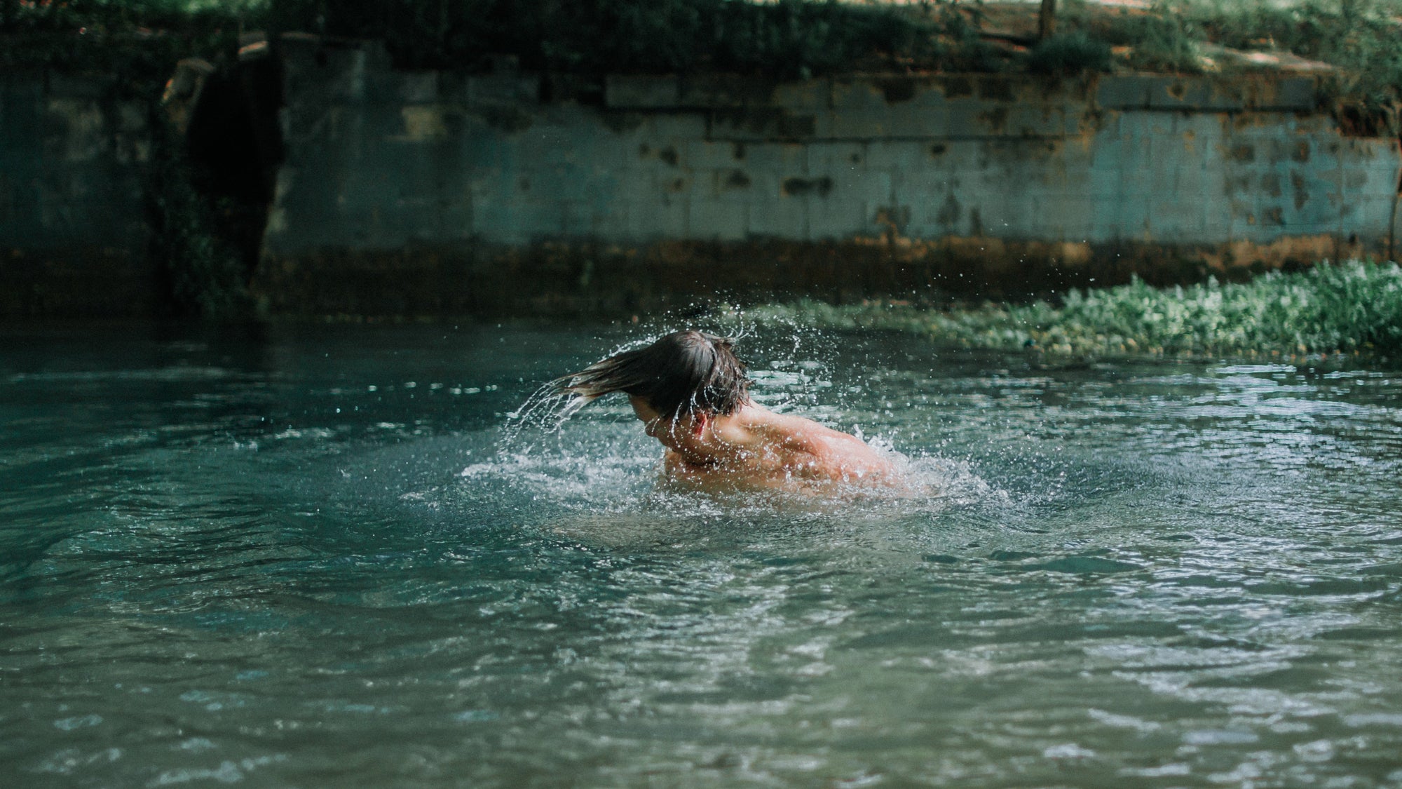 Person flipping wet hair while swimming in a green river with a mossy concrete embankment in the background