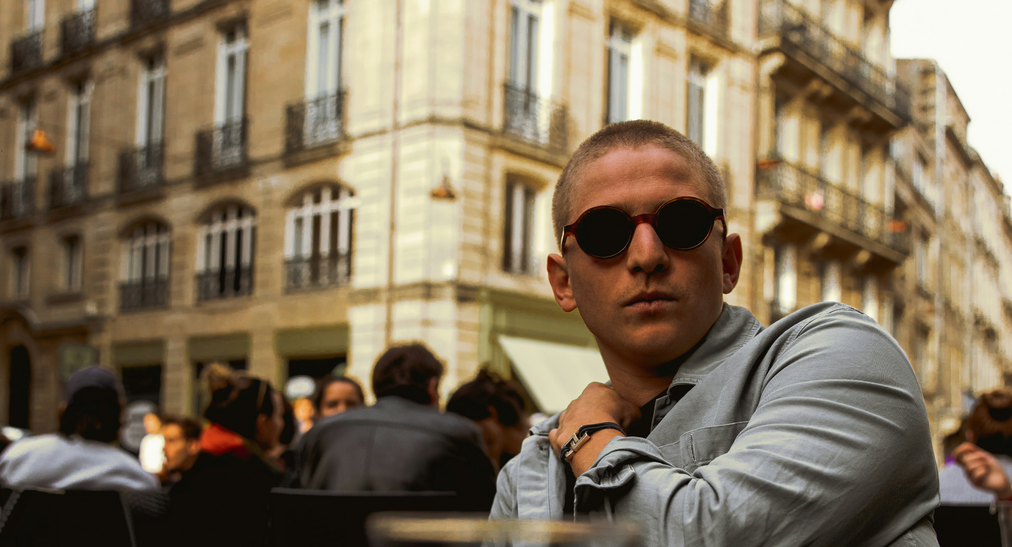 Young man with a buzzcut and round sunglasses sitting at an outdoor café, looking at the camera with blurred historic buildings and a crowd behind him