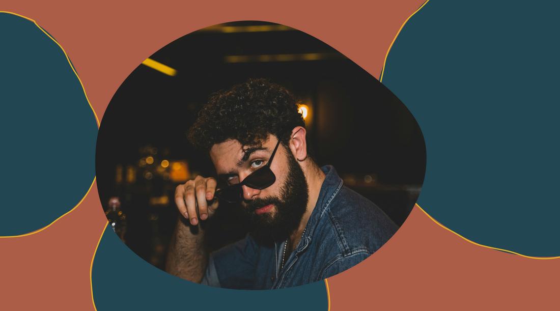 Bearded man in denim jacket lowering sunglasses, looking at camera in a dimly lit bar