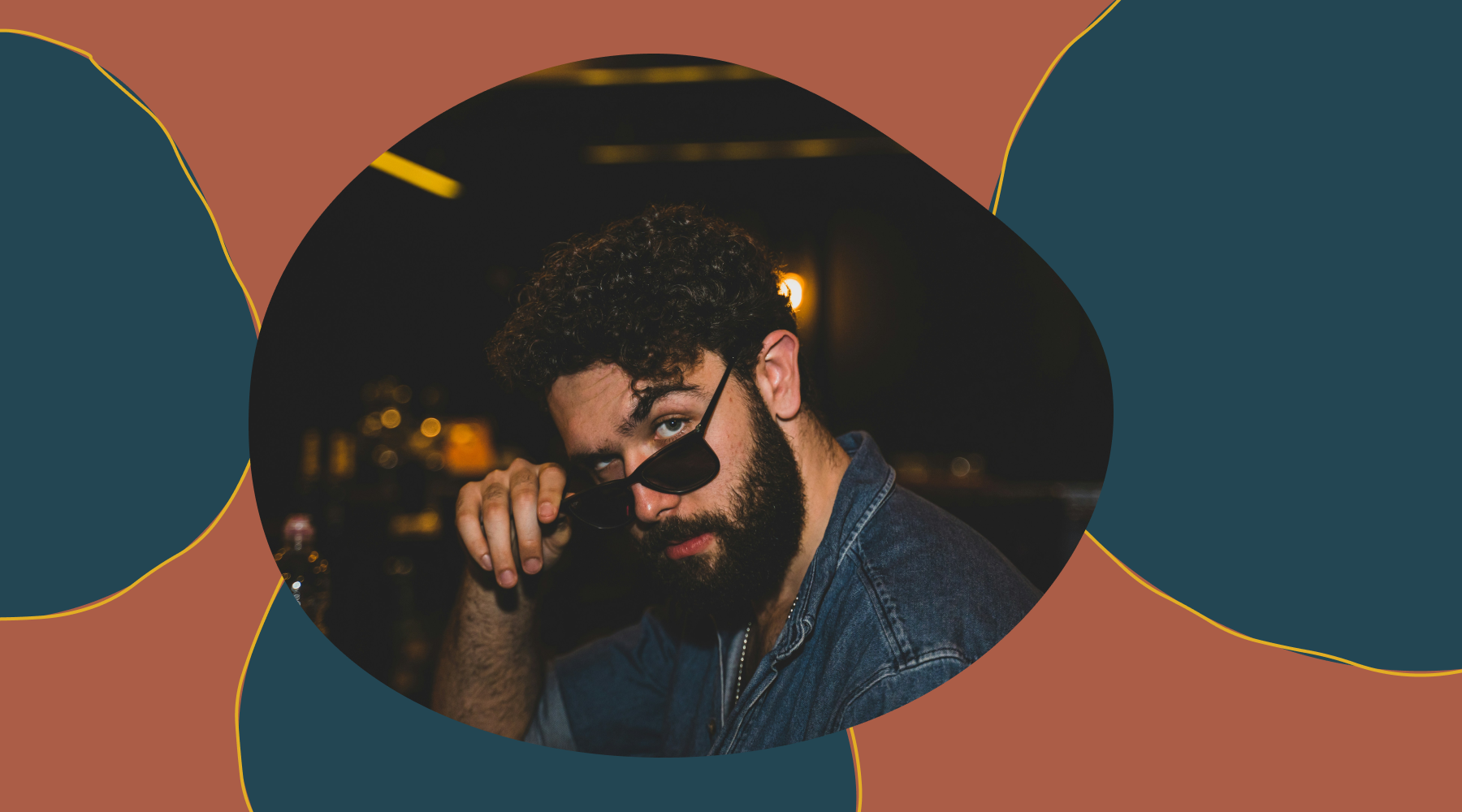 Bearded man in denim jacket lowering sunglasses, looking at camera in a dimly lit bar