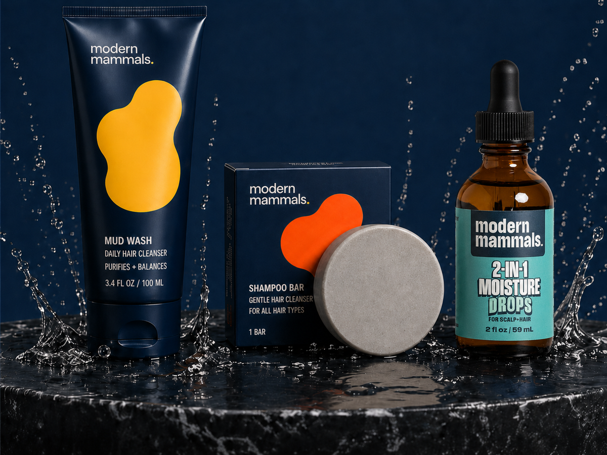 Front view of Modern Mammals products showcasing Mud Wash tube, shampoo bar, and 2-in-1 Moisture Drops on wet marble with water splashes