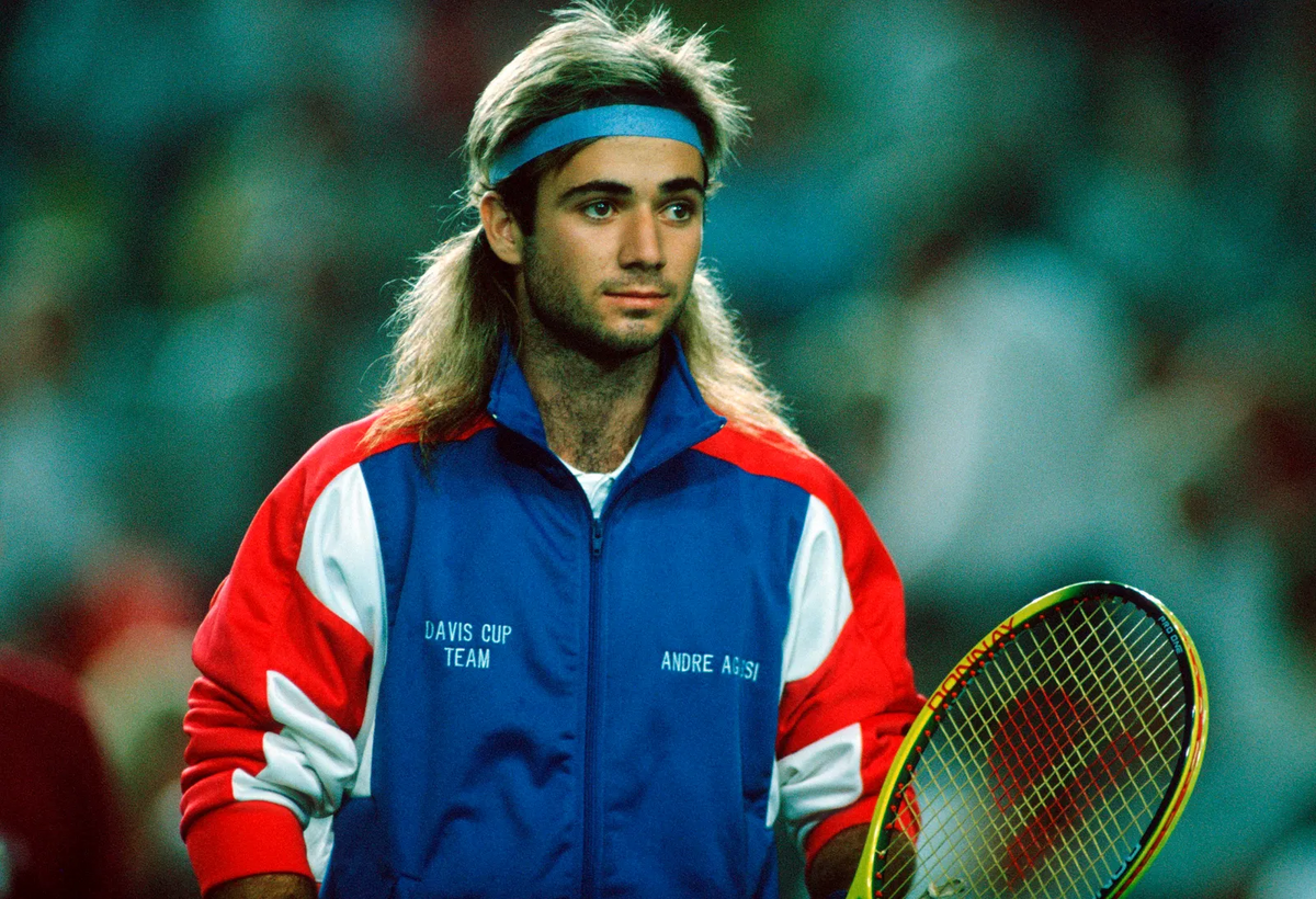 Man in retro tennis outfit holding a racket at an indoor match, wearing a blue headband and red-and-blue jacket