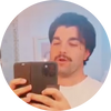 Young man taking a mirror selfie in a bathroom mirror, holding a smartphone; dark hair and mustache.