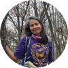 Smiling woman wearing a purple T-shirt and backpack standing among leafless trees.